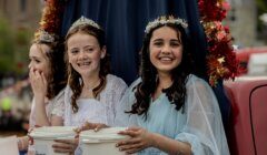 Three girls wearing dresses and tiaras sit with decorated background, smiling and holding white donation buckets.