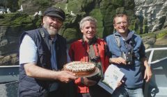 Dunter III skipper Jonathan Wills (left) presents Swiss tourist Andy Baumgartner with “The Birds of Shetland” book and cake to celebrate the 30,000th passenger with Seabirds-and-Seals, accompanied at the Noss gannetry with guide Jon Dunn (right) of Shetland Wildlife.