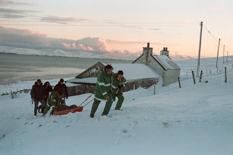 Leading ambulance paramedic (titles at the time) Peter Smith pushing a basket stretcher with a patient while with paramedic Davy Peterson, on the right, and probationer ambulance technician Michael Leask pulling the stretcher up the slope near Scarvister, at Wester Skeld. Also in the photo are family and neighbours of the patient.