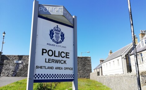 A sign outside a stone building reads "Police Scotland, Police Lerwick, Shetland Area Office" on a clear day with a blue sky.