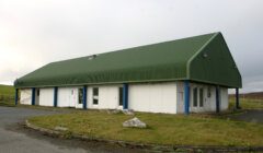 Green-roofed industrial building with multiple doors and an asphalt driveway, under an overcast sky.