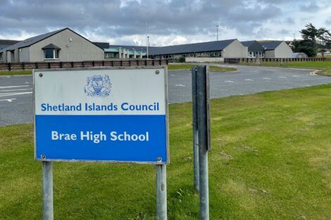 Sign reading "Shetland Islands Council Brae High School" in front of school buildings with cloudy sky in the background.