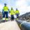Three workers in high-visibility clothing and helmets walk on a wooden path next to a large underground cable in a trench at a construction site.