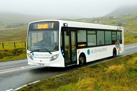 A white bus with "LERWICK E" displayed on the front LED screen is driving on a wet countryside road, with misty hills and scattered houses in the background.