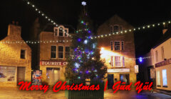 A decorated Christmas tree with colorful lights stands in a town square. The words "Merry Christmas" and "Güd Yül" are written in red across the image.