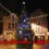 A decorated Christmas tree with colorful lights stands in a town square. The words "Merry Christmas" and "Güd Yül" are written in red across the image.