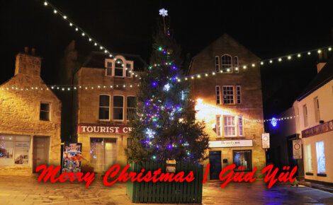 A decorated Christmas tree with colorful lights stands in a town square. The words "Merry Christmas" and "Güd Yül" are written in red across the image.