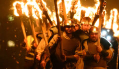 A group of people, dressed in historical attire and helmets, march at night holding large, flaming torches above their heads.