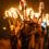 A group of people, dressed in historical attire and helmets, march at night holding large, flaming torches above their heads.