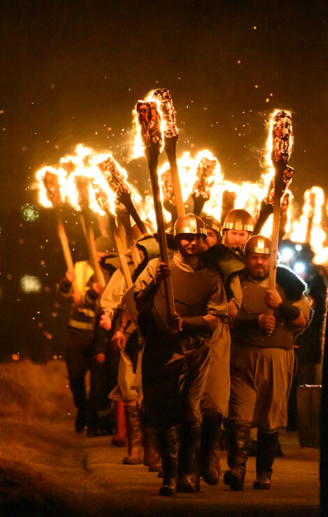 A group of people, dressed in historical attire and helmets, march at night holding large, flaming torches above their heads.