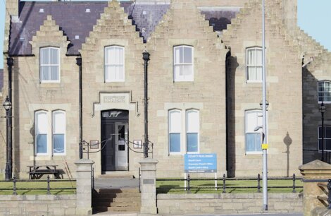 A stone building with a sign reading "Police" above the entrance, features gabled roofs and windows. A blue sign at the front indicates "County Buildings." There are benches and a lamppost nearby.