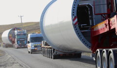 Three trucks transporting large cylindrical structures on a road in daylight, with a hilly backdrop.