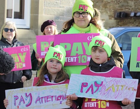 A group of people, including two children, are holding signs that read "Pay Attention" during a protest. They are wearing bright clothing and beanies with similar slogans.