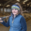 A person wearing a headband and padded vest stands in a barn holding a pitchfork over their shoulder, with sheep in the background.