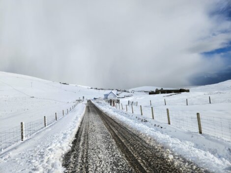 A snow-covered road flanked by fences leads to distant houses under a cloudy sky in a rural, hilly landscape.