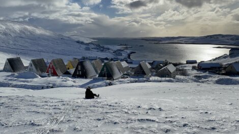 Snow-covered village with A-frame houses by a watery expanse under a cloudy sky; a person sits on the snow in the foreground.