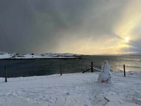 Snow-covered landscape with a snowman near a fence by a body of water under a cloudy sky. Small buildings and snowy hills are visible in the background.