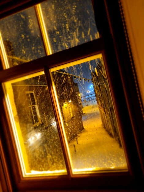 View through a frosty window showing a dimly lit snow-covered alley at night, illuminated by warm yellow light and distant blue lighting.