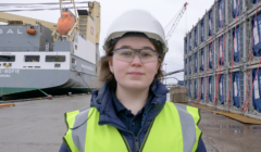 A person wearing a hard hat, safety glasses, and a high-visibility vest stands on a dock in front of a cargo ship and stacked containers.