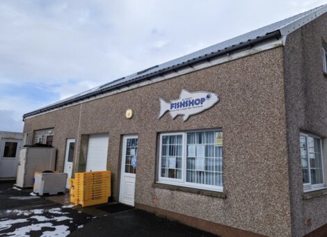 A small, beige building with a sign in the shape of a fish that reads "Fish Shop." Stacks of yellow crates are outside near the entrance. The sky above is partly cloudy.