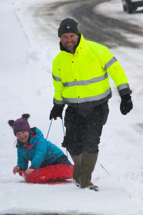 An adult in a bright yellow jacket pulls a child on a red sled through the snow on a snowy day. The child smiles while sitting on the sled.