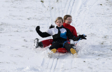 Two children wearing winter jackets and gloves are sledding down a snowy hill on a red sled. They appear to be enjoying the ride.