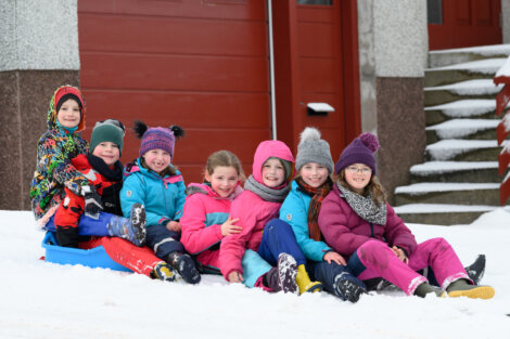 A group of children dressed in winter clothing sit together on the snow, smiling and enjoying the snowy day.