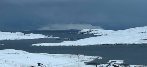 Snow-covered landscape with a body of water and small islands in the foreground. A dark, cloudy sky looms above the distant mountains.