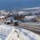 Snow-covered suburban street with several vehicles driving, surrounded by houses, and a mix of cloudy and blue sky in the background.
