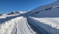 A snow-covered road winds through a hilly landscape with tire tracks visible, flanked by snow walls. A clear blue sky stretches above.