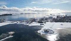 Aerial view of a coastal town surrounded by snow and ice, with a calm body of water and a snow-covered island in the background under a partly cloudy sky.