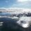 Aerial view of a coastal town surrounded by snow and ice, with a calm body of water and a snow-covered island in the background under a partly cloudy sky.