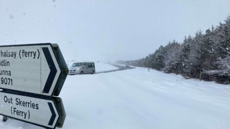 Snow-covered road with a vehicle driving in the distance. Road signs pointing to Whalsay and Out Skerries ferry directions are visible in the foreground. Snowfall obscures visibility.
