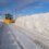 A snowplow clears a path through a thick layer of snow, with snow banks piled high on both sides of the road under a blue sky with clouds.