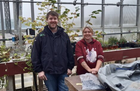 Two people stand inside a greenhouse, smiling at the camera. The person on the left wears a dark jacket, and the person on the right wears a red hoodie. There are plants and equipment around them.