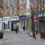 A street scene in a small town with colorful bunting flags overhead. People walk past various shops, and a "No Entry" sign is visible in the foreground.