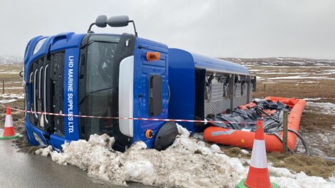 A blue truck labeled "LHD Marine Supplies Ltd." lies overturned on a snow-covered roadside next to several orange cones and a red barrier tape.