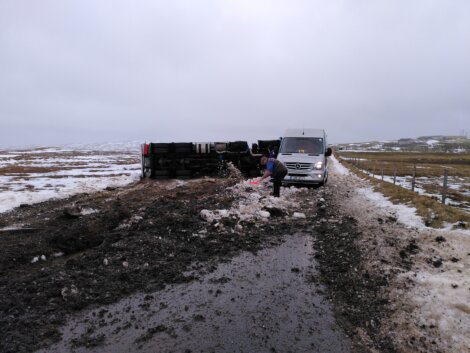 A person removes debris from a snowy, muddy road next to an overturned truck and a white van on a cloudy day.