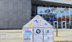 A small, see-through structure with various information panels stands in front of a large building with colorful art above the entrance.