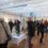 A group of people, some with coats and bags, view artwork and socialize inside a bright gallery space with wooden floors and white walls. A table with snacks and drinks is in the center.