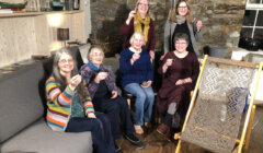 A group of six women, smiling and holding up small glasses, are gathered in a cozy room with wooden and stone walls. A loom is visible in the foreground.