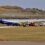 A small passenger plane with "Eastern Airways" livery is stationed on a runway, surrounded by several utility vehicles and personnel, with a grassy landscape and rocky hills in the background.