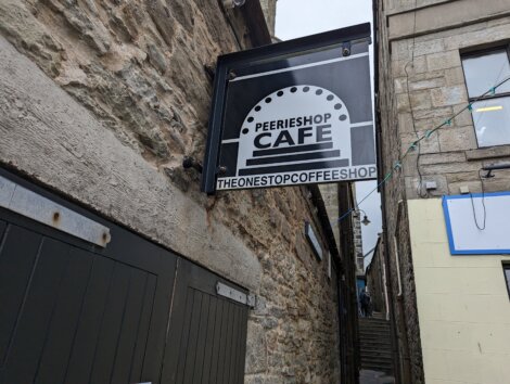 Sign for PeerieShop Café, labeled "The One Stop Coffee Shop," mounted on a stone building along a narrow alley.