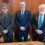 Three men in suits stand in a row in a wood-paneled room. The man on the left has gray hair and glasses, the man in the middle has dark hair and is smiling, and the man on the right has white hair and a beard.