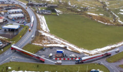 Aerial view of a large truck transporting an oversized wind turbine blade through a roundabout in a rural area with patches of snow on the ground.