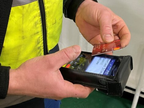 Person in a yellow jacket holds a POS terminal, swiping a red contactless card to make a payment.