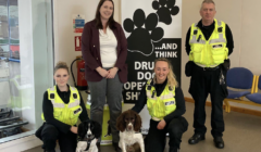 Four police officers, three wearing reflective yellow vests, pose with two dogs in front of a banner that reads "Paws...and think" and "Drug dogs operate here.