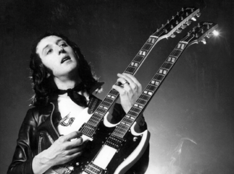 A person wearing a leather jacket plays a double-neck electric guitar.