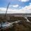 A crane and construction materials are present at a wind farm development site in a hilly and remote area, with several wind turbine components visible near a dirt road winding through the landscape.