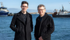 Two men in dark jackets stand by the water, with fishing boats docked in the background on a clear day.